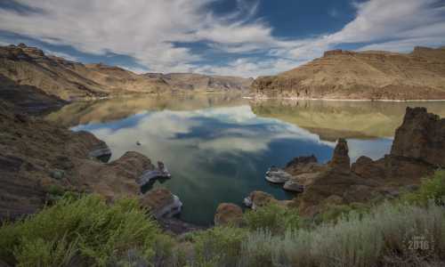 A serene landscape featuring a calm lake surrounded by rocky hills and blue skies with fluffy clouds.