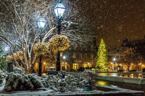 A snowy night scene featuring a lit Christmas tree, glowing street lamps, and festive decorations in a charming plaza.