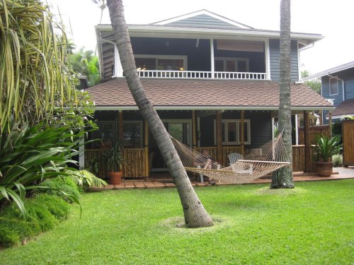 A charming two-story house with a porch, surrounded by lush greenery and a hammock in the yard.