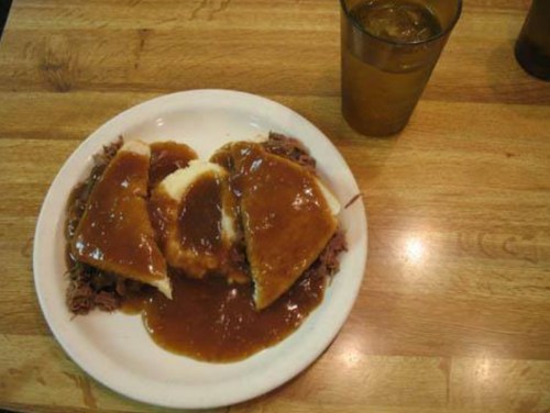 A plate of sliced meat with gravy, mashed potatoes, and a glass of iced tea on a wooden table.
