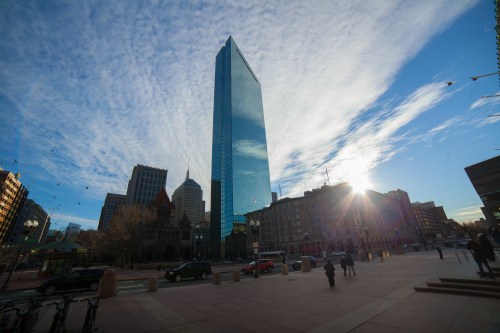A tall glass skyscraper reflects the sky, surrounded by city buildings and a clear blue atmosphere.