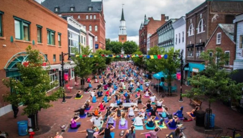Aerial view of a large group practicing yoga in a vibrant, urban street lined with trees and colorful decorations.
