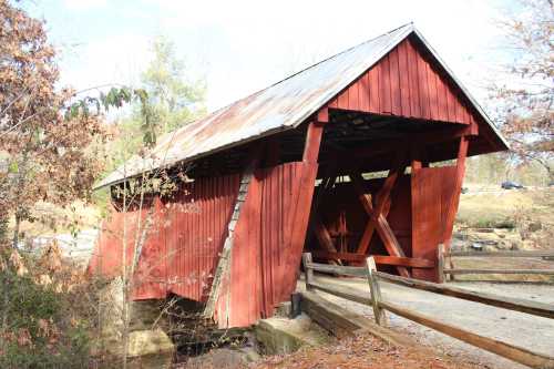 A red covered bridge with a metal roof, surrounded by trees and a gravel path, over a small stream.