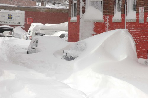 A car is buried in deep snow beside a red brick building, with snowdrifts covering the street and nearby vehicles.