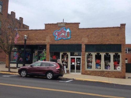 A brick storefront with colorful signage and large windows, featuring a parked car in front and an American flag.