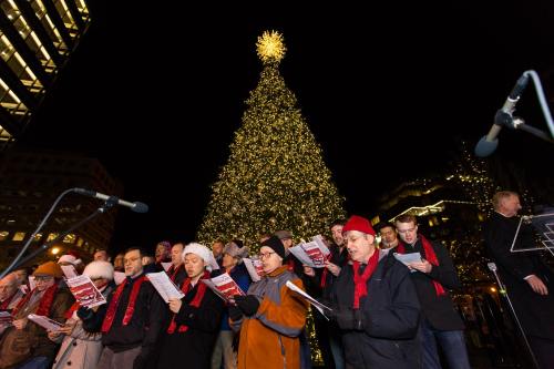 A choir sings in front of a brightly lit Christmas tree at night, wearing festive hats and scarves.