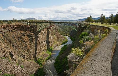 A green bridge spans a deep canyon with a winding river below, surrounded by trees and rocky terrain under a cloudy sky.