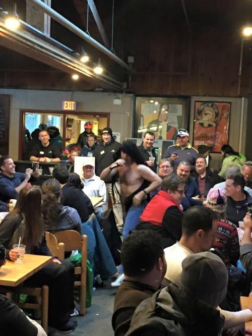 A lively bar scene with a shirtless performer singing, surrounded by an engaged audience enjoying drinks and laughter.