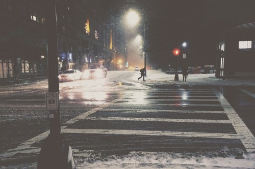 A snowy street at night, with streetlights illuminating a crosswalk and a few pedestrians in the distance.