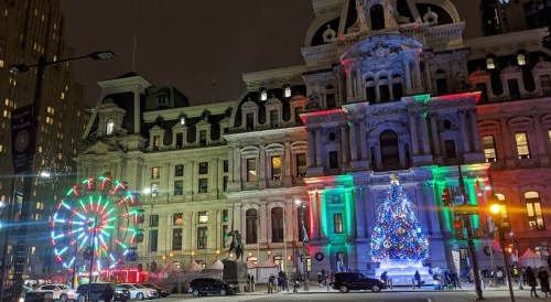 A festive city hall adorned with colorful lights, a large Christmas tree, and a nearby illuminated Ferris wheel at night.