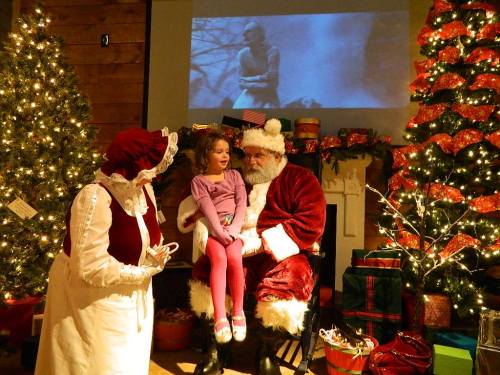 A girl sits on Santa's lap, smiling, while a woman in a festive dress stands nearby, surrounded by Christmas decorations.