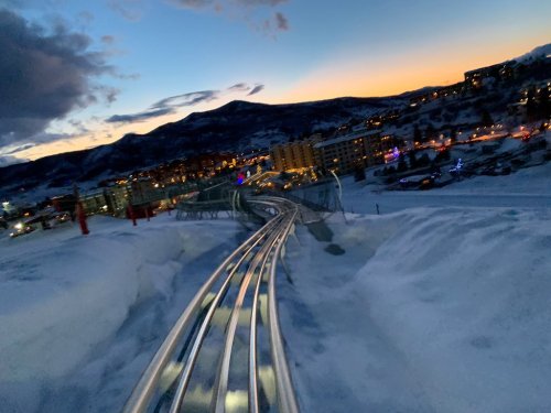 A snowy landscape with a winding roller coaster track, illuminated buildings, and a sunset in the background.
