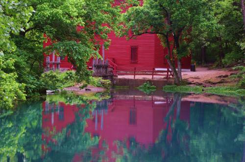 A red building reflected in calm water, surrounded by lush green trees.