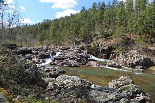 A serene river flows over rocks, surrounded by lush green trees and a clear blue sky with scattered clouds.