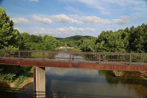 A metal bridge spans a calm river, surrounded by lush green trees and rolling hills under a partly cloudy sky.