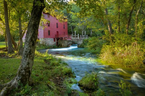 A red mill beside a flowing stream, surrounded by lush green trees and grass in a serene natural setting.