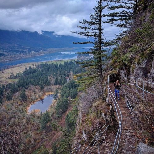 A winding path along a cliff with a view of a river and forested valley under a cloudy sky.