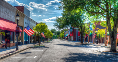 A quiet street lined with trees and shops, featuring American flags and a clear blue sky.