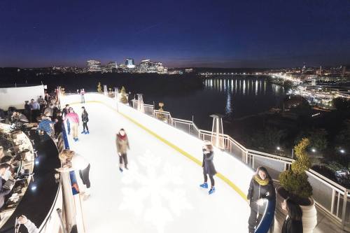People ice skating on a rooftop rink at night, with a city skyline and river in the background.
