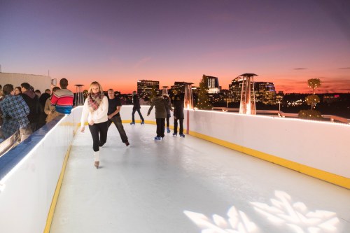 A rooftop ice skating rink at sunset, with people skating and city buildings in the background.