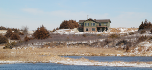 A house on a hill overlooking a calm lake, surrounded by dry grass and sparse trees under a clear blue sky.