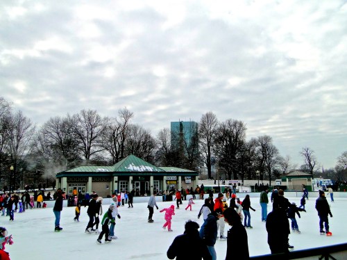 A busy outdoor ice skating rink with people of all ages skating, surrounded by trees and a cloudy sky.