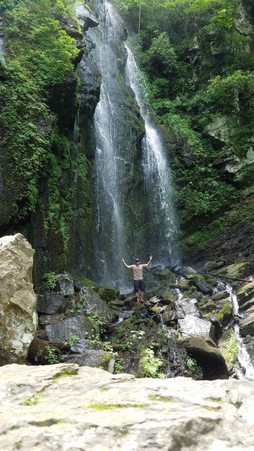A person stands with arms raised in front of a lush waterfall surrounded by rocks and greenery.