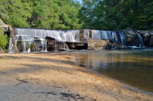 A serene waterfall cascades over rocks into a calm pool, surrounded by lush greenery and sandy shore.