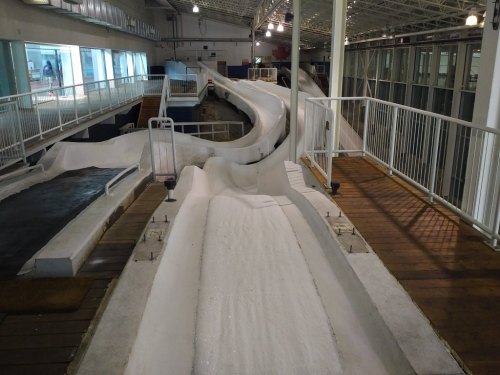 A view of a winding indoor water slide with white surfaces and wooden decking, surrounded by railings and bright lighting.