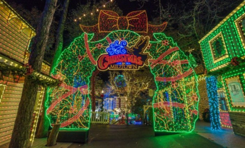 Colorful holiday lights decorate an archway reading "Christmas Midtown," surrounded by festive buildings and trees.