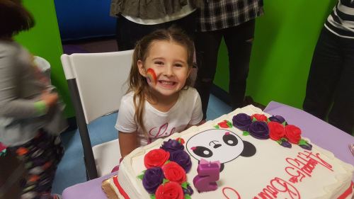 A smiling girl with a panda-themed cake, colorful face paint, and flowers around the cake at a celebration.
