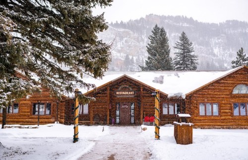 A cozy log cabin restaurant surrounded by snow-covered trees and mountains in a winter landscape.