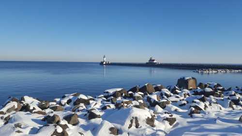 A snowy shoreline with large rocks, leading to a calm lake and distant lighthouses under a clear blue sky.