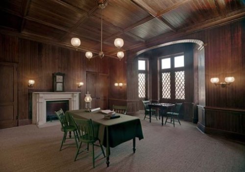 A vintage room with wooden walls, a fireplace, a table, and green chairs, illuminated by hanging lights.