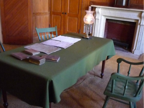 A green table with books and papers, a lamp, and wooden chairs in a room with a fireplace and wooden walls.