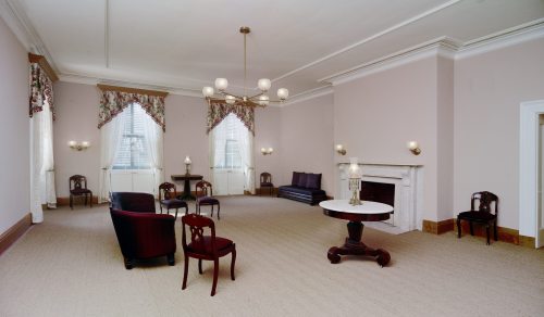 A spacious, elegantly furnished room with a fireplace, chandelier, and seating arranged around a central table.