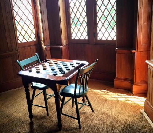 A wooden checkers table with two chairs, illuminated by sunlight through diamond-patterned windows.
