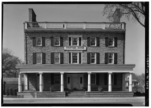 Historic stone building with a sign reading "Frontier House," featuring a porch and multiple windows.