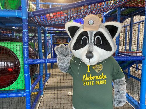 A raccoon mascot wearing a cap and a "I ❤️ Nebraska State Parks" shirt waves in a colorful play area.
