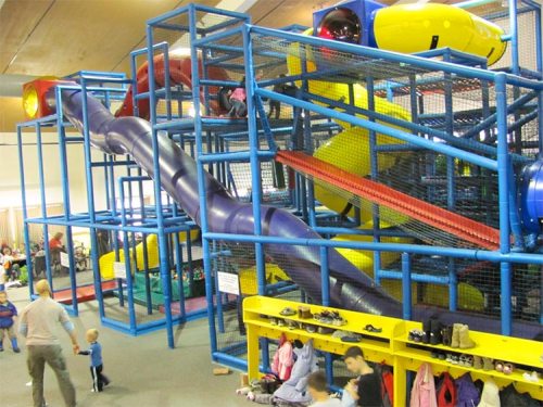 Colorful indoor playground structure with slides and climbing areas, filled with children and parents. Shoes are lined up below.