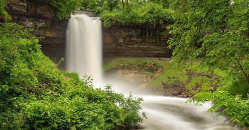 A serene waterfall cascades over rocks, surrounded by lush green foliage and flowing water below.