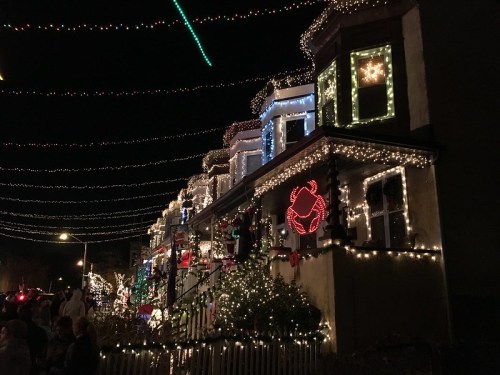 A festive street scene at night, featuring houses adorned with colorful Christmas lights and decorations.