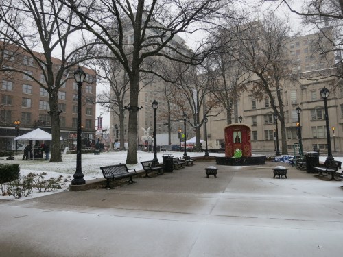 A snowy park scene with benches, trees, and a small gazebo, surrounded by buildings and a festive setup in the background.