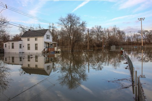A flooded house surrounded by water, reflecting in the still surface, with trees and power lines in the background.