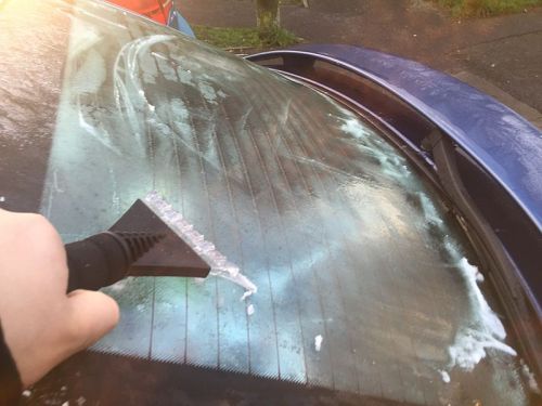 A hand using a scraper to remove ice and snow from a car's rear windshield on a sunny day.