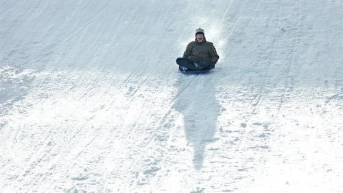 A person sledding down a snowy hill, sitting on a sled with a joyful expression.