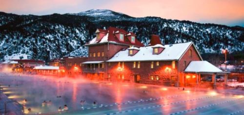 A scenic view of a historic building by a steaming outdoor pool, surrounded by snow-covered mountains at sunset.