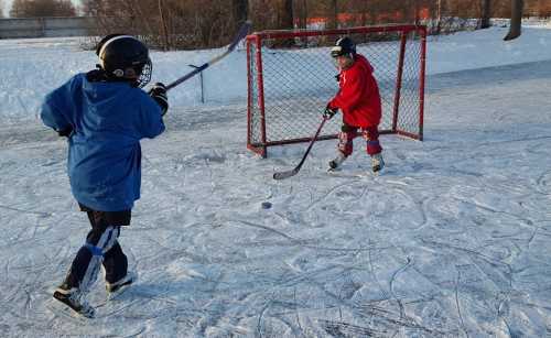 Two children playing ice hockey on a frozen rink, one shooting a puck towards a red goal while wearing helmets and gear.