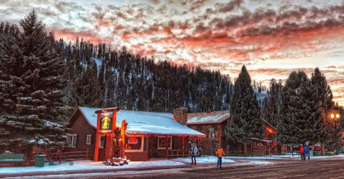 A snowy mountain town at sunset, featuring rustic buildings, evergreen trees, and people walking along the street.
