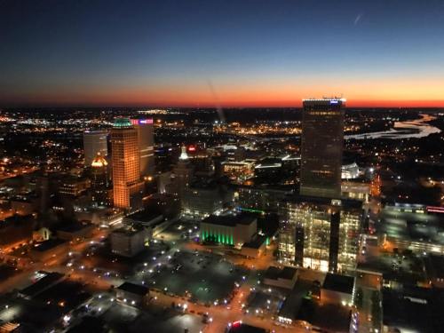 Aerial view of a city skyline at sunset, with illuminated buildings and a river in the background.